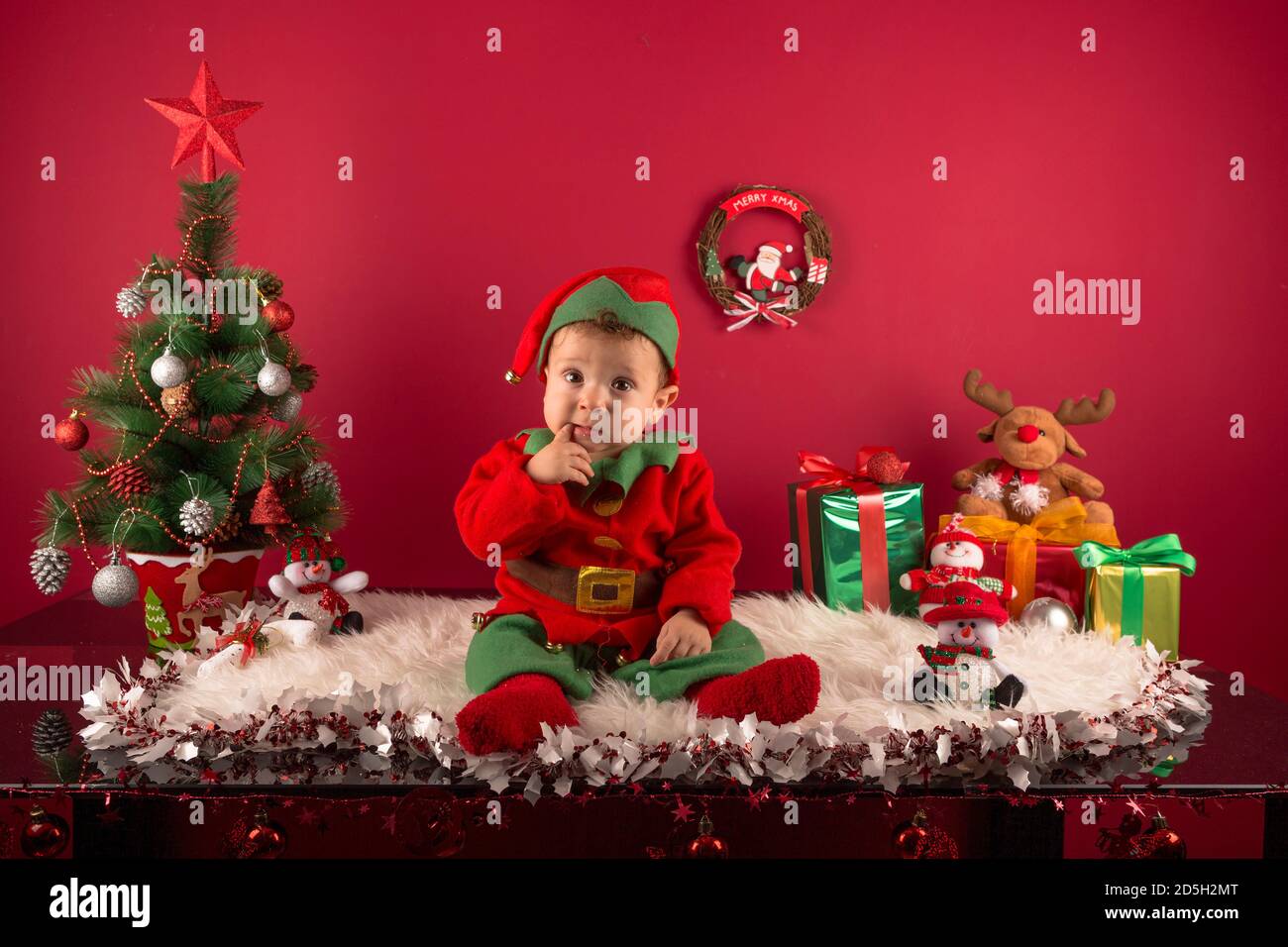 Sweet one-year-old boy dressed as an elf, in Christmas decoration, with gifts and typical tree Stock Photo