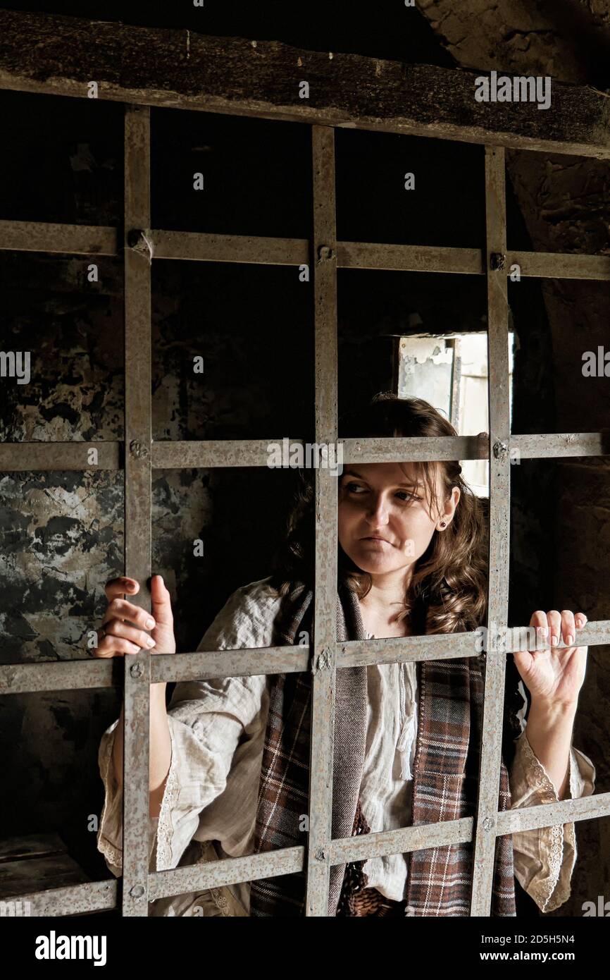 A young woman in a vintage dress from the 18-19 century looks through the bars of a prison cell. Stock Photo