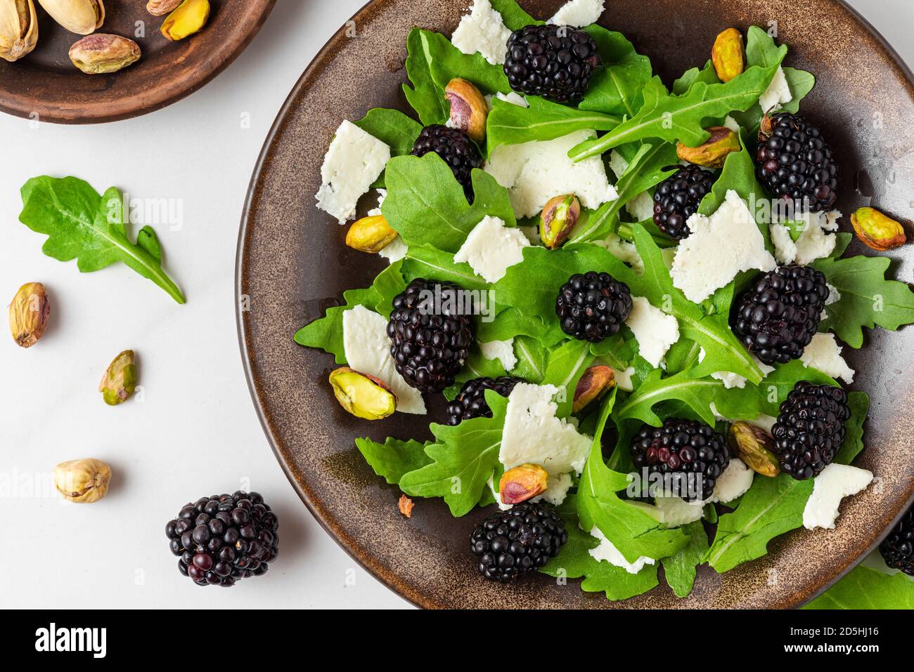 Fresh salad with arugula, goat cheese, blackberries and pistachios in a plate on white background. top view. healthy diet food Stock Photo