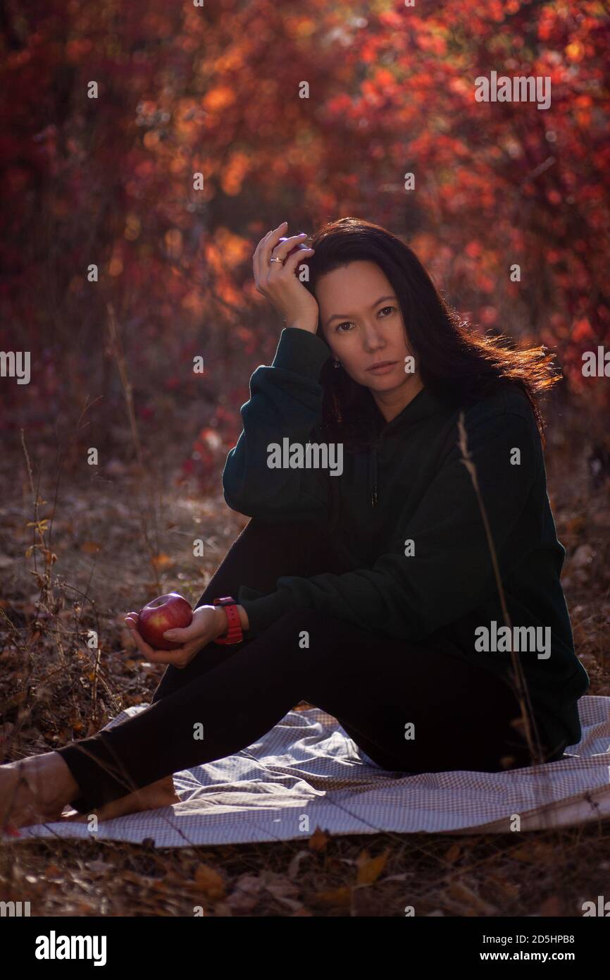 beautiful Asian young woman sitting on a blanket and holding apples in the autumn forest at sunset Stock Photo