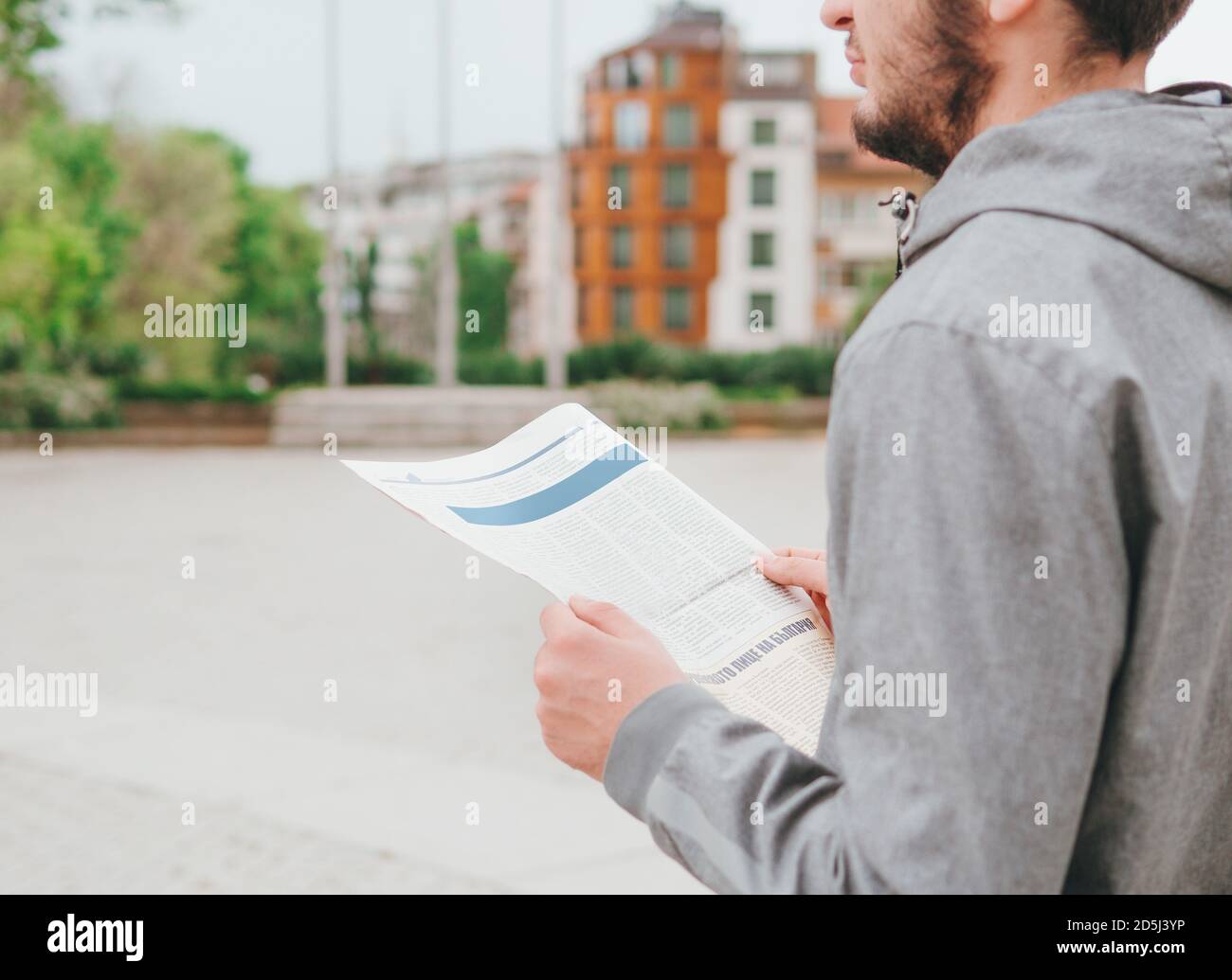 Hands of a man in gray hoodie holding a newspaper looking away to the side thoughtful, daydreaming isolated on city street background Stock Photo