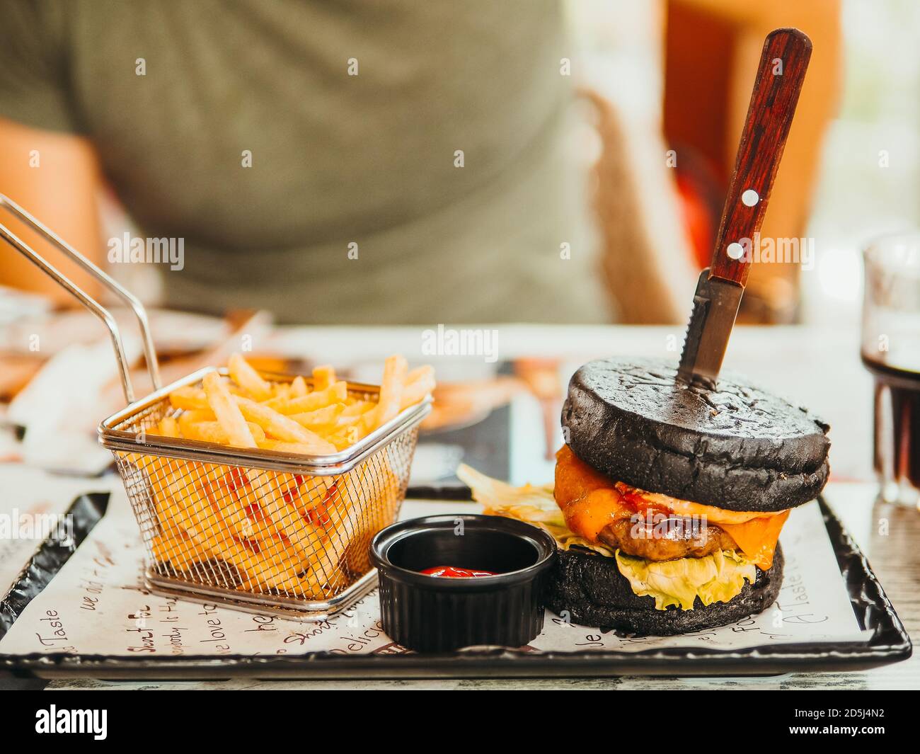 Unhealthy eating and fast food concept. Junk food, black burger and french fried potatoes in front of man silhouette on a table in a restaurant. View Stock Photo