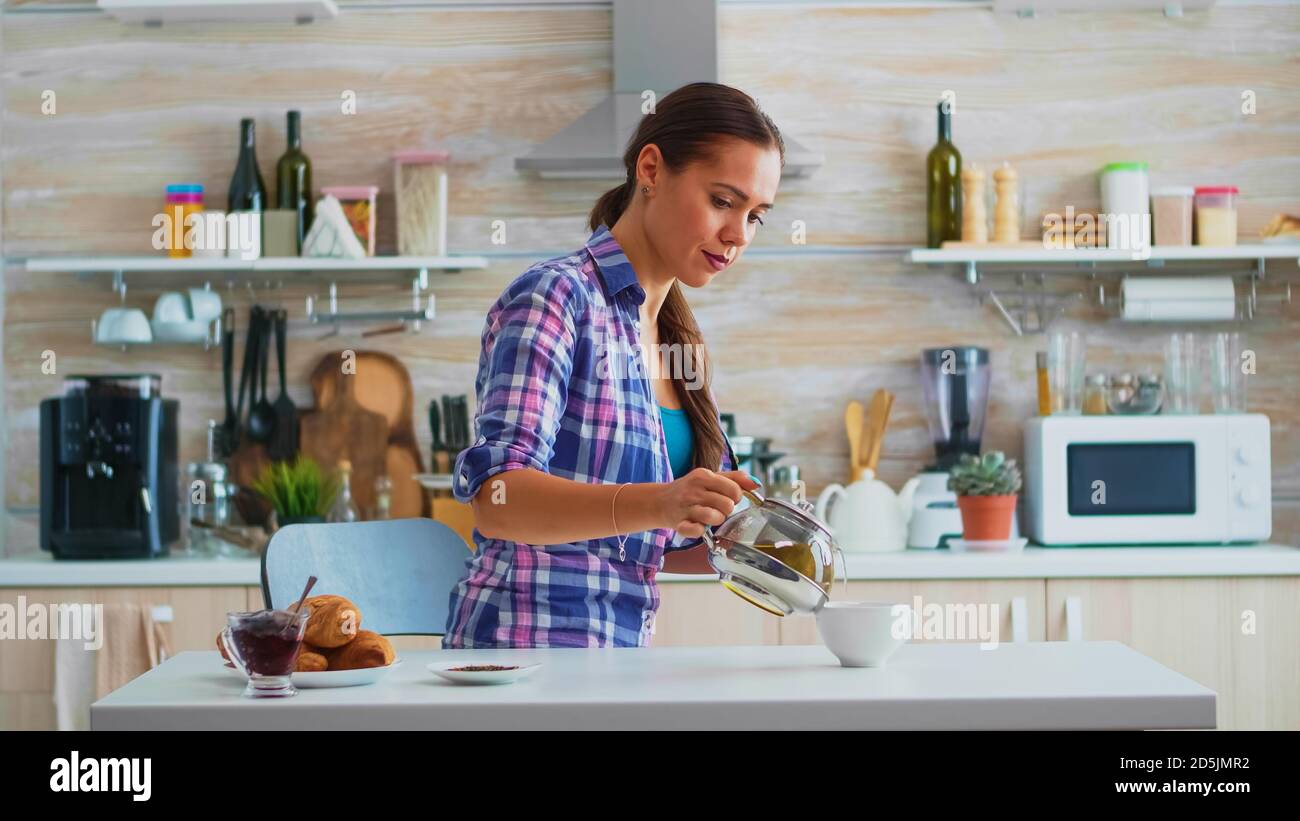 Housewife Pouring Hot Green Tea In Cup In The Morning Sitting In The Kitchen Woman Ejoying A Healthy Herbal Tea At Breakfast Using Teacup And Teapot Sitting Near The Table Holding The