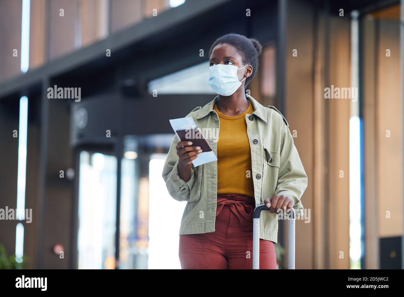 African young woman in protective mask with luggage holding tickets while going along the airport Stock Photo