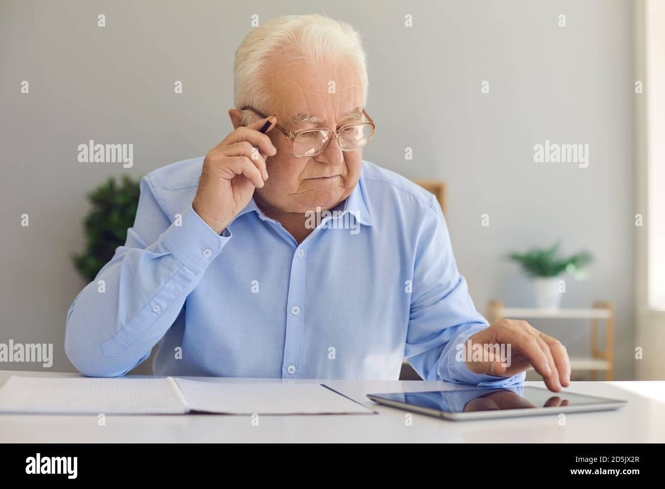 Serious old man sitting at desk at home using tablet computer and taking notes in notebook Stock Photo