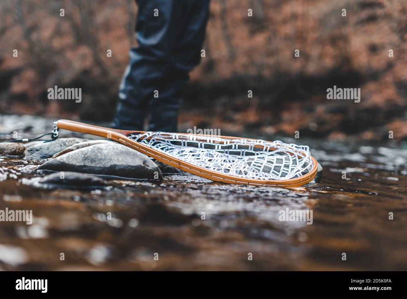 Fishing landing net on rocks in river with blured fisherman behind. Stock Photo