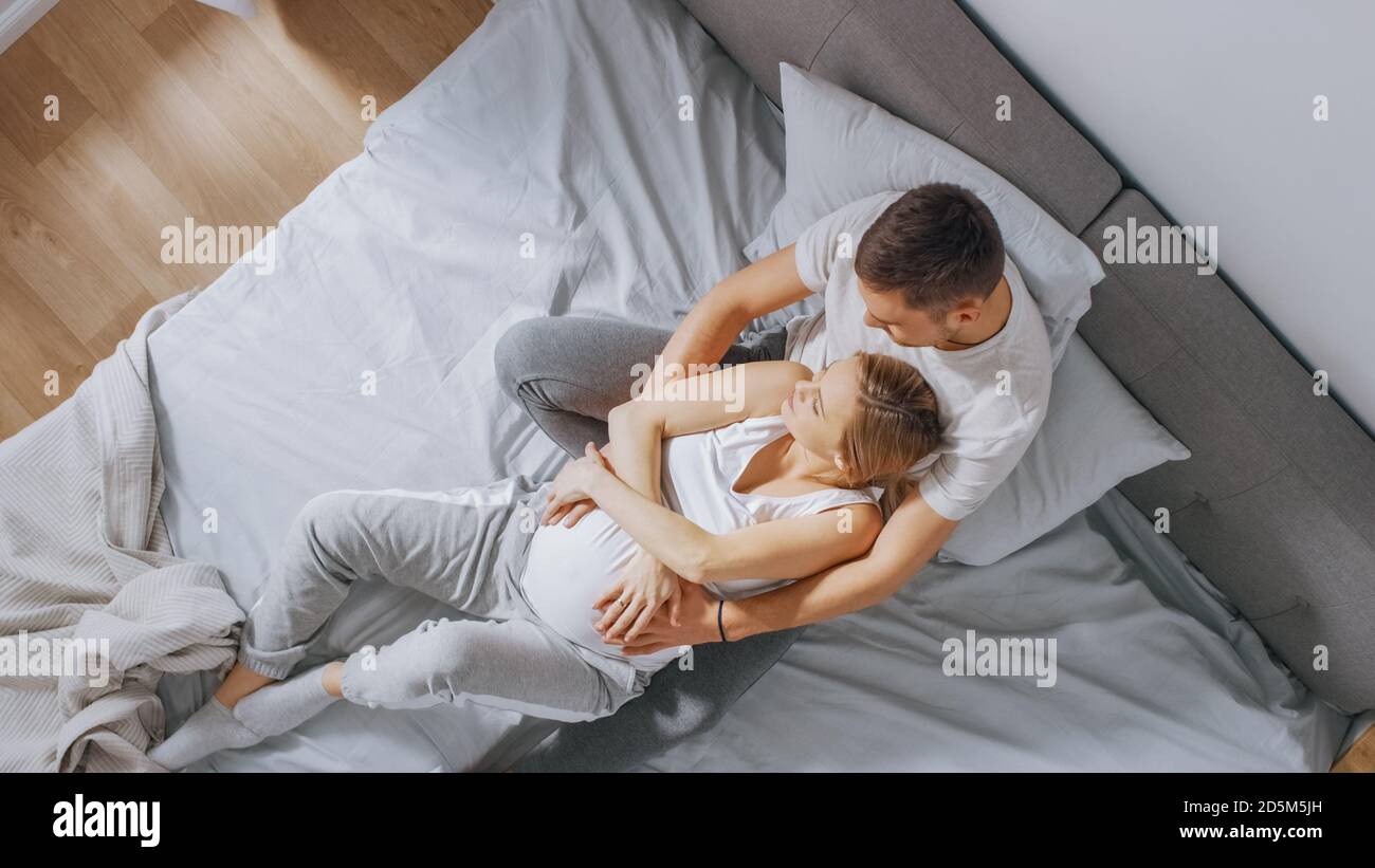 Happy Young Couple Cuddling Together in the Bed, Young Woman is Pregnant and Loving Husband Holds Her In Embrace and Caresses Her Belly Tenderly Stock Photo