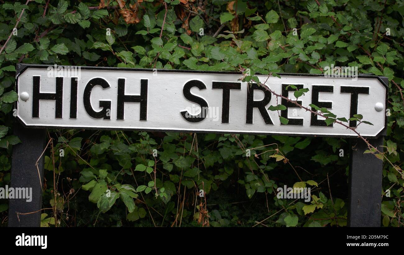 Old traditional English high street sign in small rural village surrounded by dark green foliage. Shops highstreet shopping town centre. Stock Photo