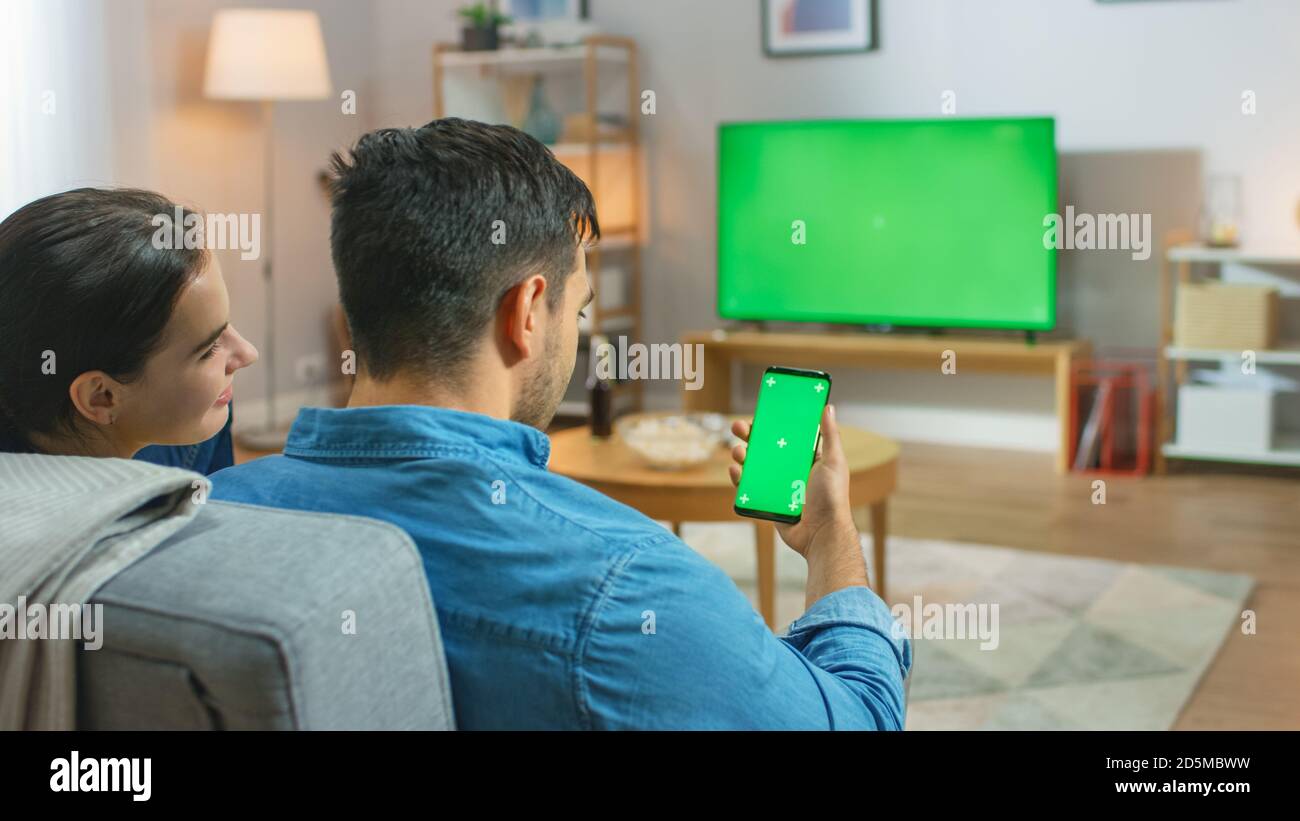 Happy Couple Sitting At Home in the Living Room Watching Green Chroma Key Screen Television, Relaxing on a Couch. Guy also Uses Green Mock-up Screen Stock Photo