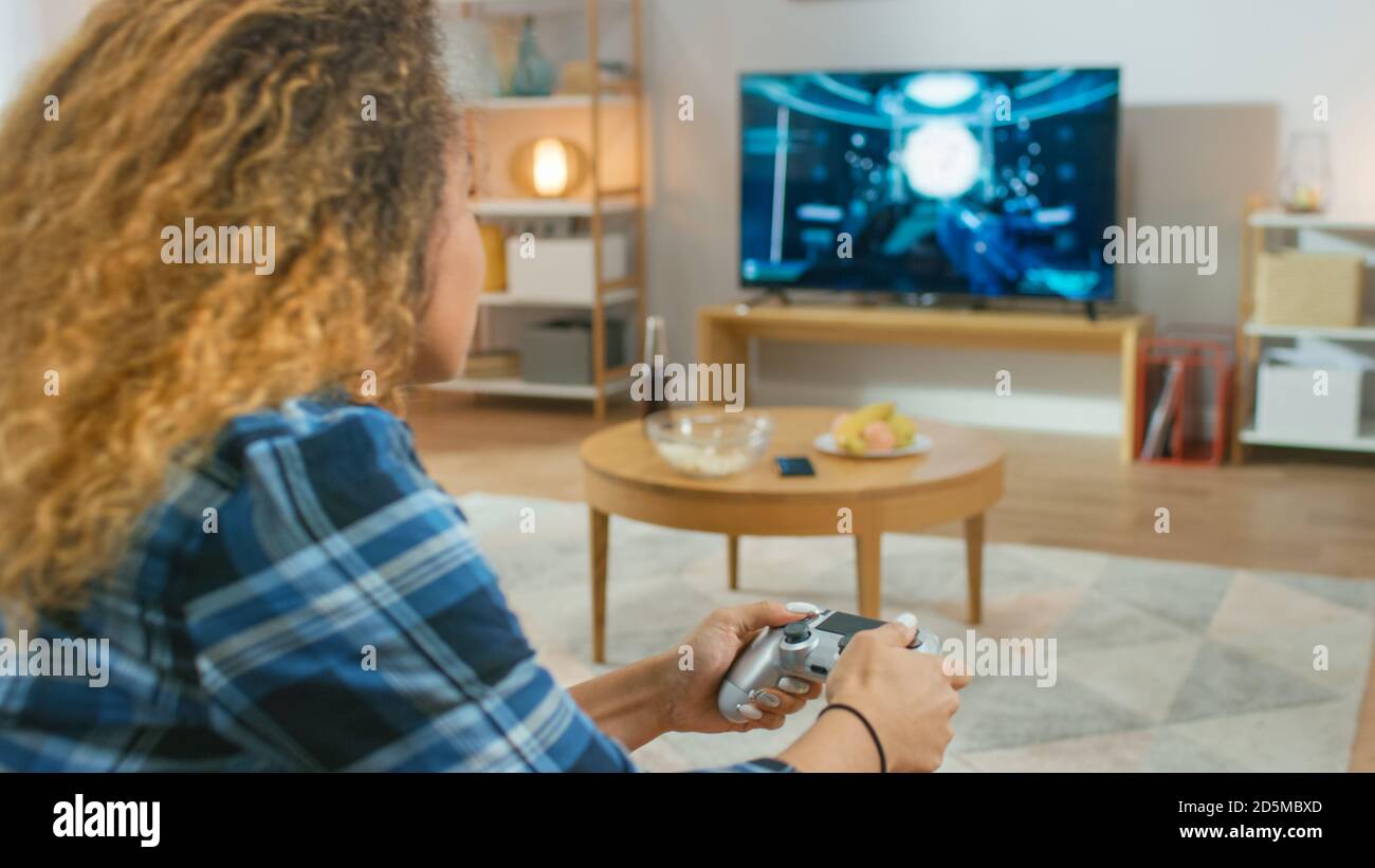 In the Living Room Girl Sitting on a Couch Holds Controller Playing in a Console Video Game, 3D Action Shooter Gameplay Shown on TV Screen. Stock Photo