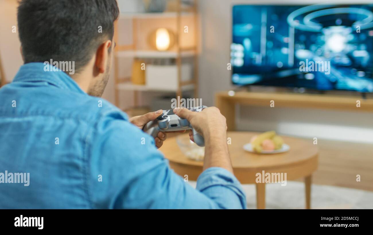 In the Living Room Man Sitting on a Couch Holds Controller Playing in a Console Video Game, 3D Action Shooter Gameplay Shown on TV Screen. Stock Photo