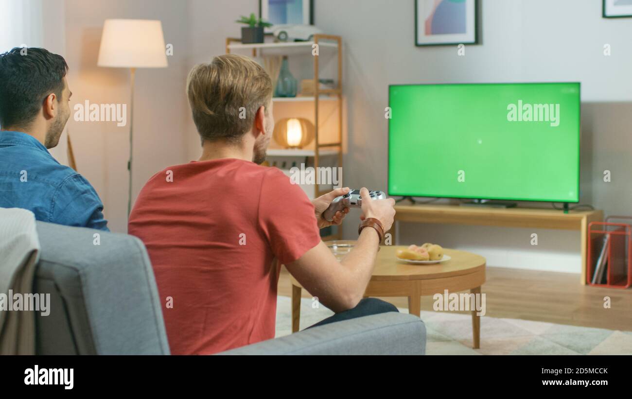 In the Living Room Two Friends Sitting on a Couch Holding Controllers Playing Competitive Video Game on a Green Chroma Key TV Screen. Stock Photo