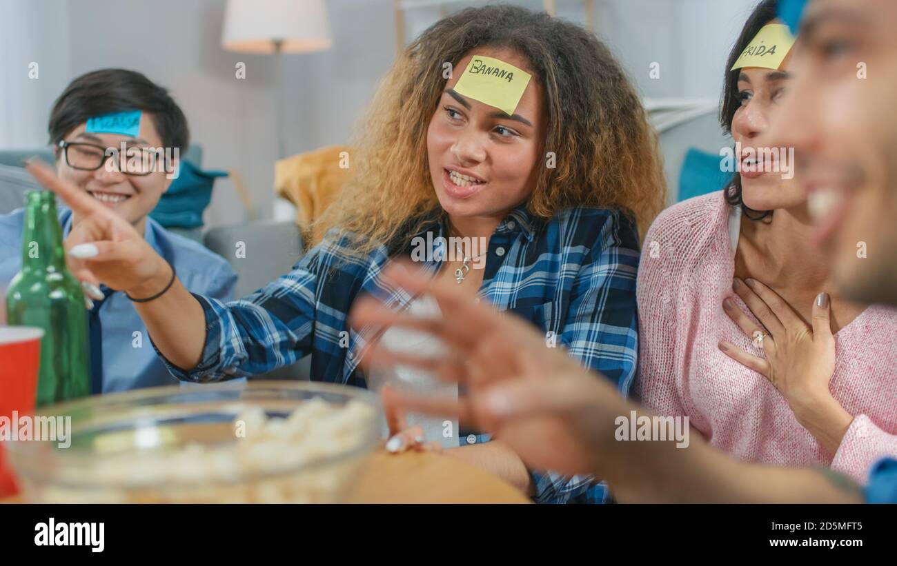 In the Living Room Diverse Group of Fun Loving Friends Playing 'Who am I' game With Sticky Papers Attached to Foreheads. Girls Points Her Finger to Stock Photo