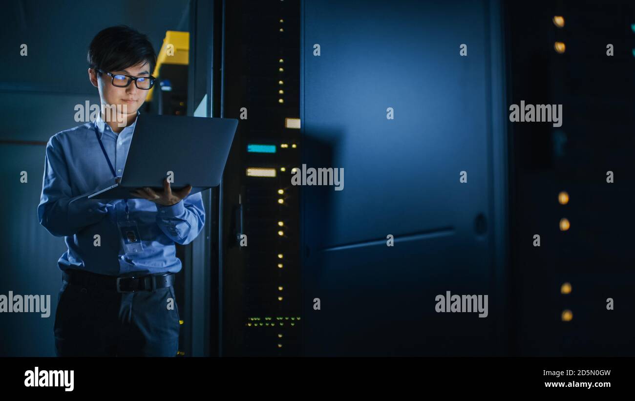 In Dark Data Center: Male IT Specialist Stands Beside the Row of Operational Server Racks, Uses Laptop for Maintenance. Concept for Cloud Computing Stock Photo