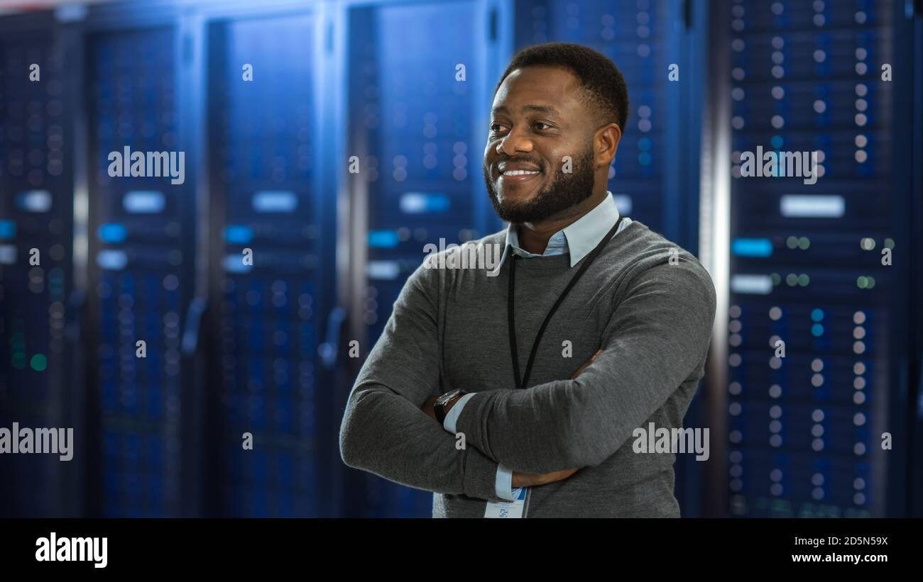 Black Data Center IT Technician Standing in the Middle of a Server Rack Corridor. He is Standing with Crossed Arms and is Smiling. Stock Photo