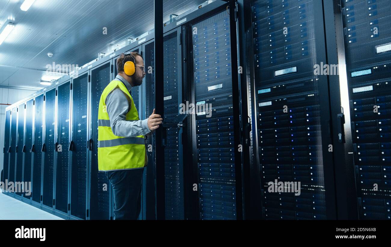 IT Administrator in High Visibility Vest is Opening the Door to the Server Rack. He's Working on Laptop in Data Center while Standing Before Them Stock Photo