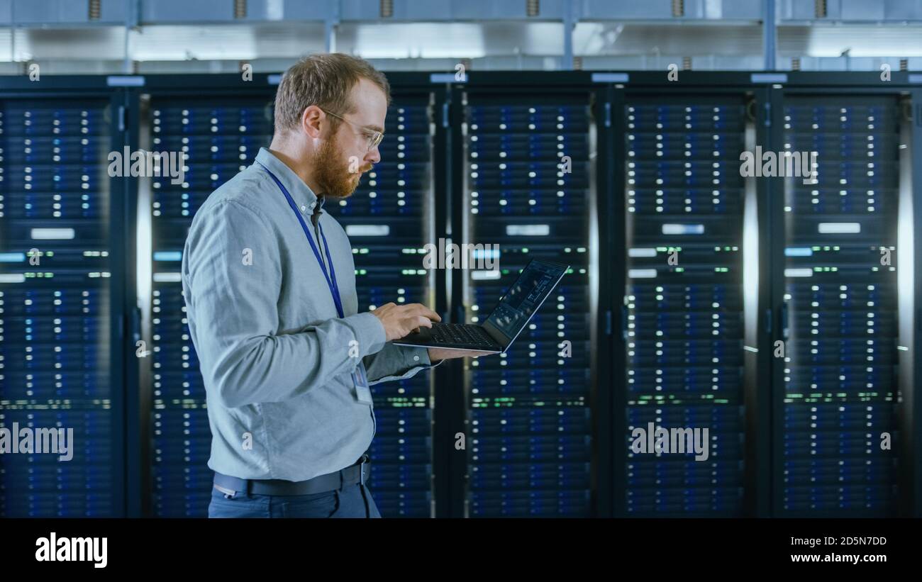 Bearded Data Center IT Engineer in Glasses is Walking Through Server Rack Corridor with a Laptop Computer. He is Visually Inspecting Working Server Stock Photo