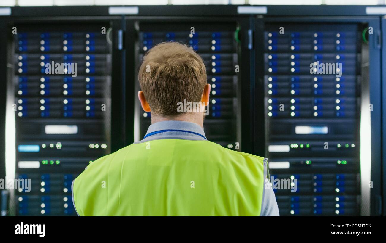 Back Shot of an IT Administrator in High Visibility Vest Walking Towards a Server Rack in Data Center Room. Stock Photo