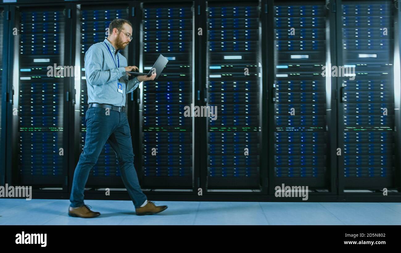Bearded Data Center IT Professional Walking Through Server Rack Corridor with a Laptop Computer. He Stops and Wirelessly Inspects Working Server Stock Photo