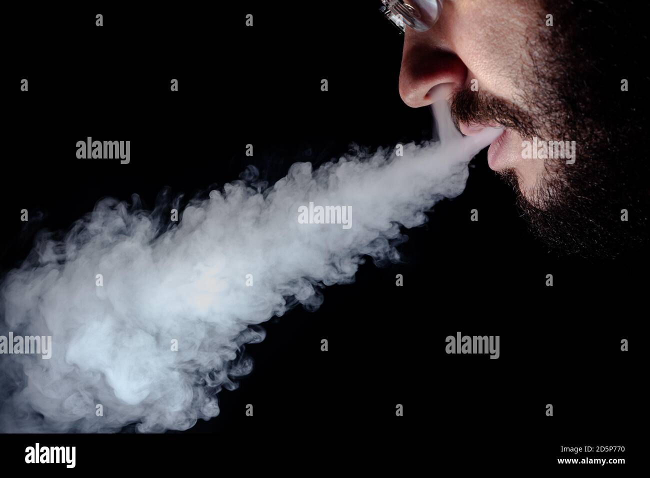 A man with a black beard and a mustache in glasses smokes a hookah and exhales a stream of smoke Stock Photo