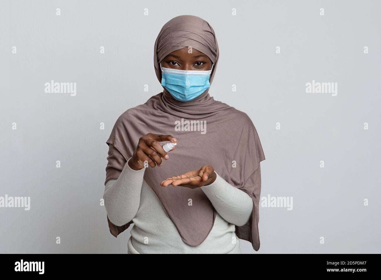 Coronavirus Precautions. Black muslim woman in mask applying disinfectant spray on hands Stock Photo