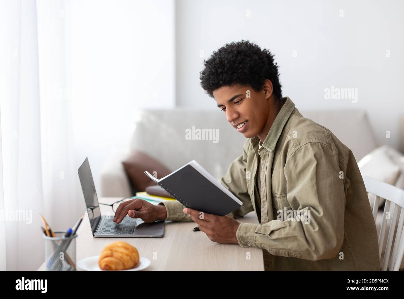 E-learning concept. Handsome black teenager using his notes to make online home assignment on laptop indoors Stock Photo
