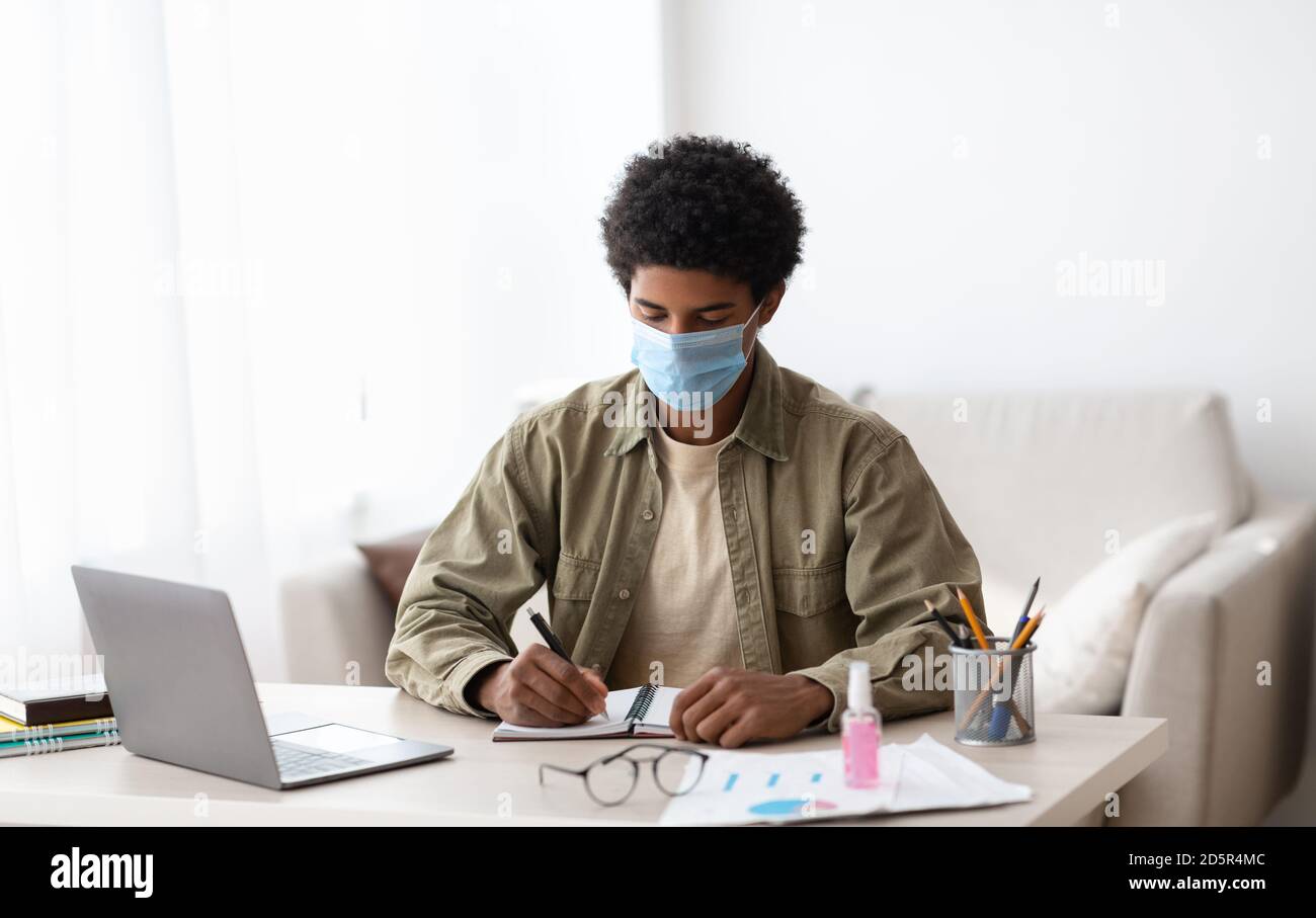 Distance education during coronavirus epidemic. Focused black guy in medical mask taking notes near laptop at home Stock Photo