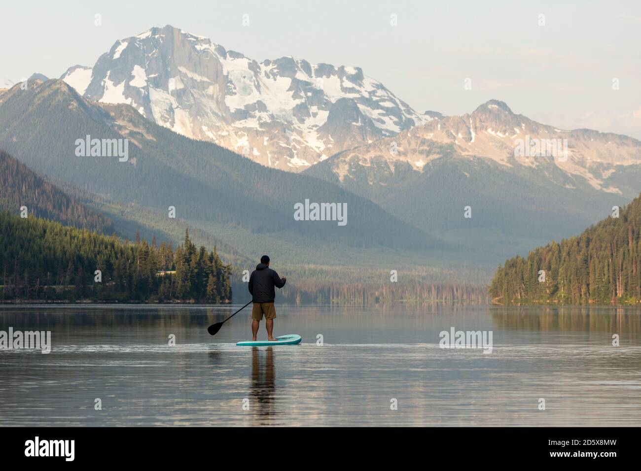 Back view of anonymous male on paddleboard on calm surface of Duffey lake and enjoying landscape of mountains in morning in British Columbia Stock Photo