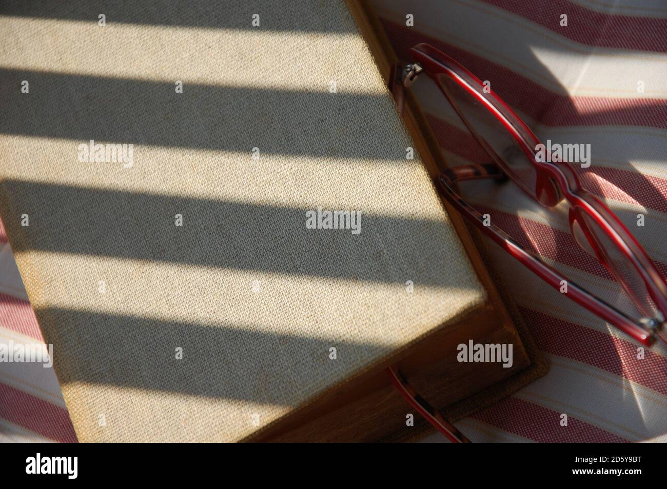 some light and shadow strips on a book cover on a chair strip cushion and red eyeglasses aside close up. summer readings. Stock Photo