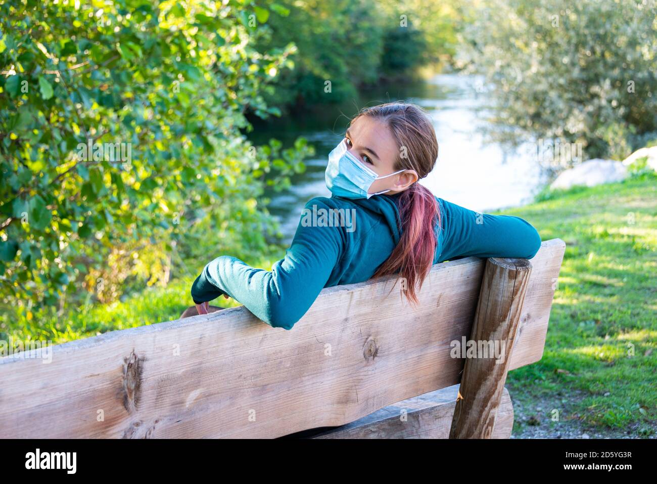 young woman sitting on park bench wearing a face mask  beside a river alone ; Stock Photo