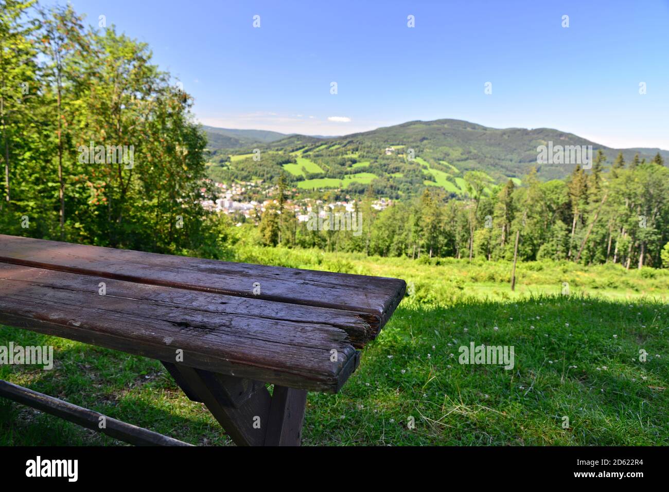 Old wooden table overlooking a mountain valley. Green meadow in the background with forest and mountains. Blue sky without clouds. Cracked old wood. S Stock Photo