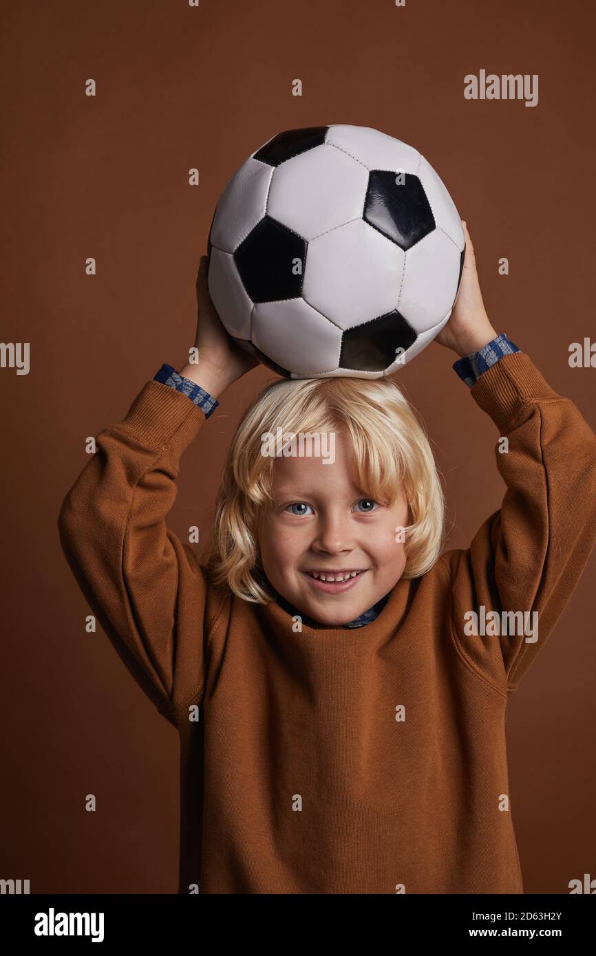 Portrait of cute child with blonde hair holding football over his head and looking at camera against the brown background Stock Photo