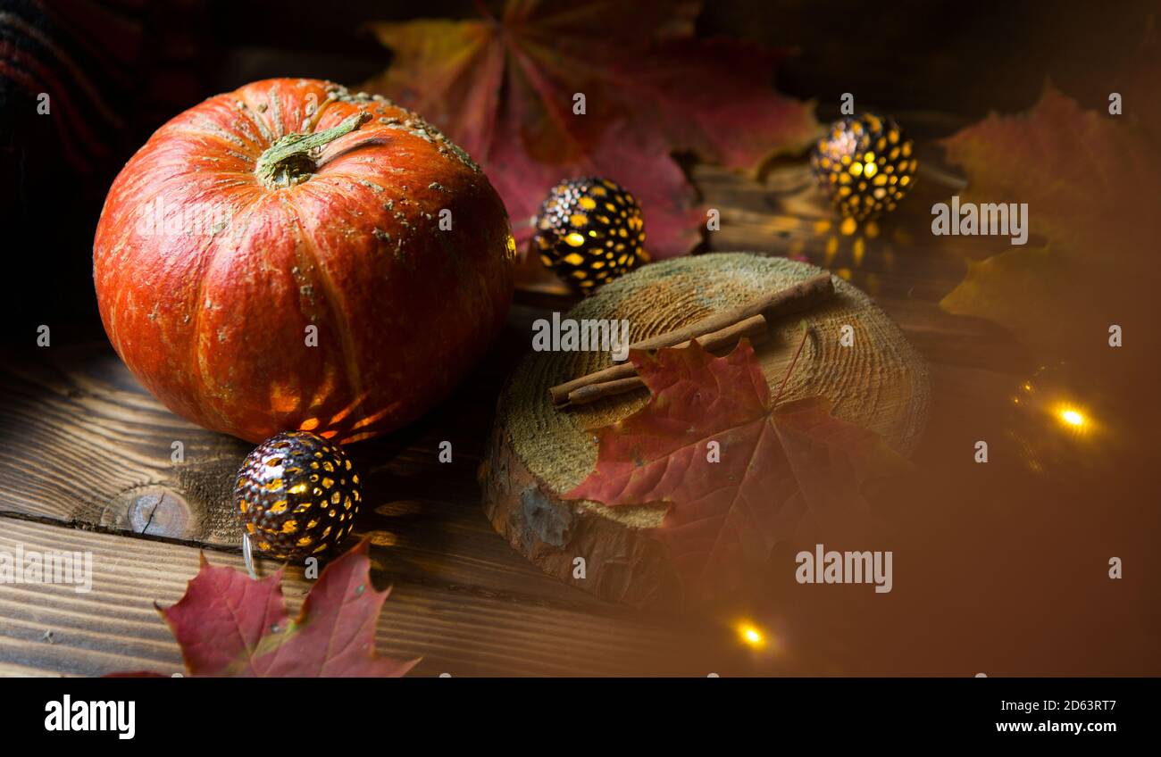 Orange natural round pumpkin on a wooden table with fallen yellow and red maple leaves, cinnamon sticks. Lights garlands, warm autumn atmosphere, than Stock Photo