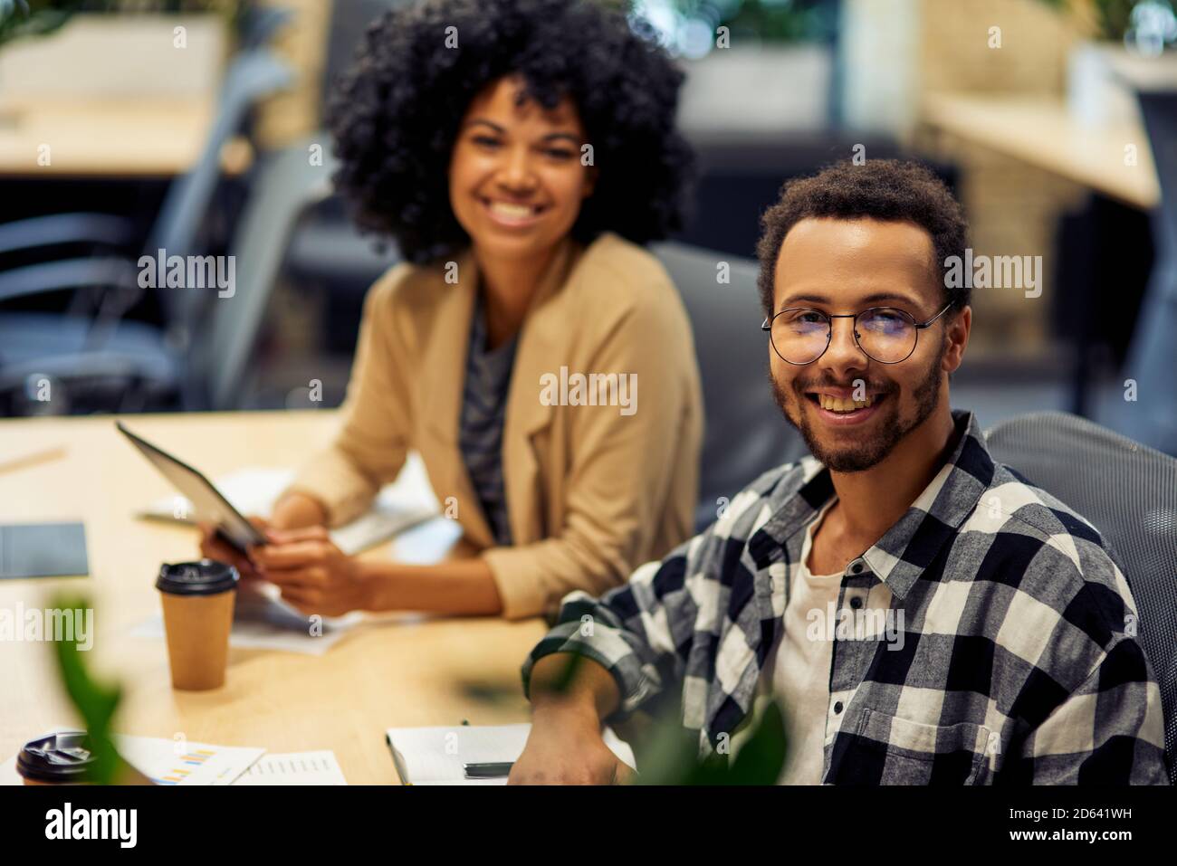 Two young cheerful male and female office workers sitting at desk , smiling at camera while working together in coworking space. Office life, teamwork and business Stock Photo