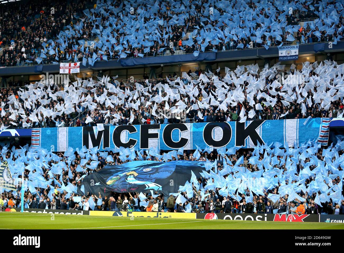 Manchester City fans wave flags ahead of kick-off near a banner that reads 'MCFC OK' Stock Photo