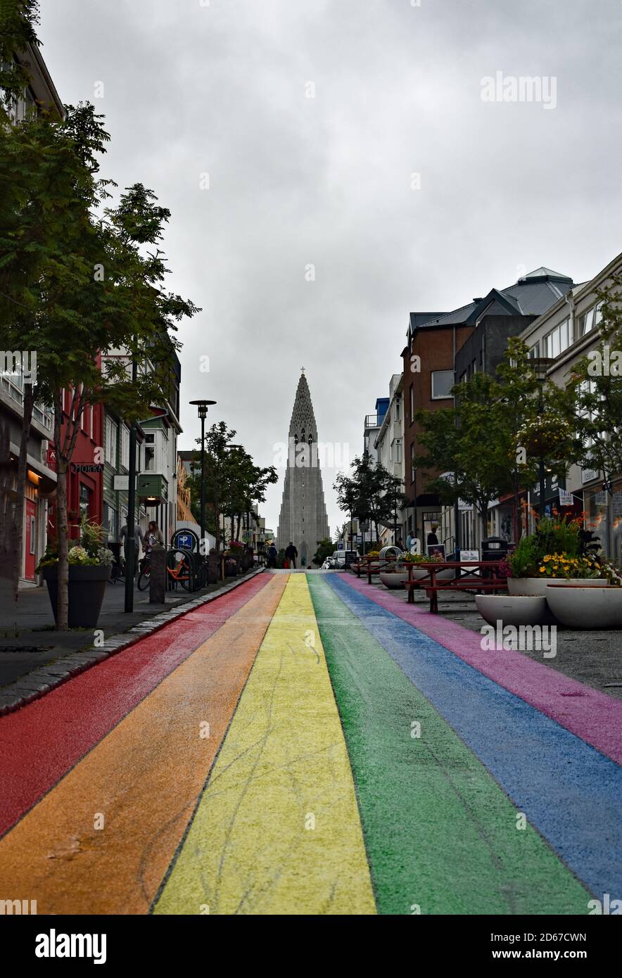 A rainbow painted road (Skolavordustigur) leads to Hallgrimskirkja Church in Reykjavik, Iceland. Trees and plants line the shop filled street. Stock Photo