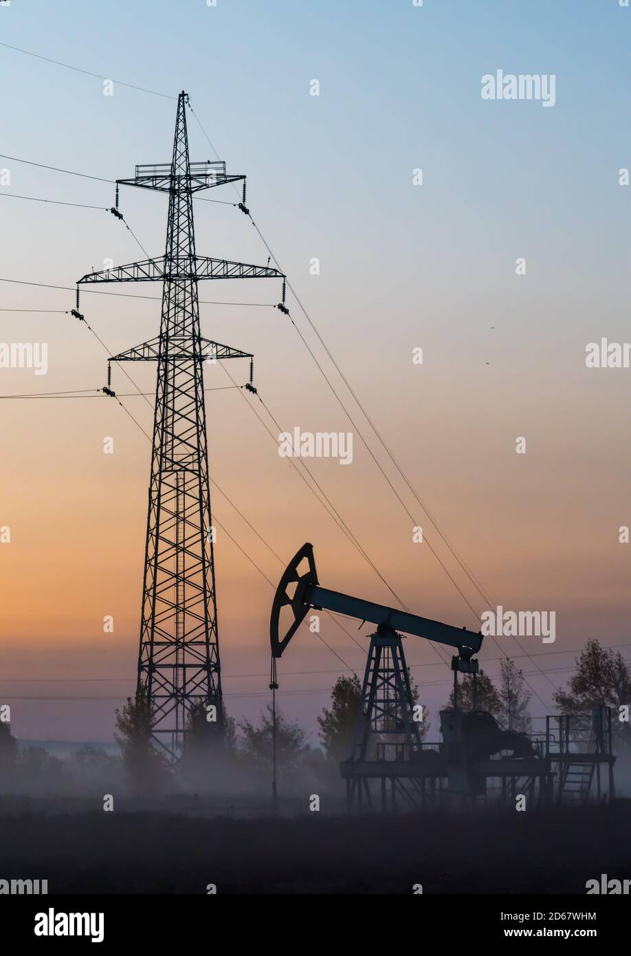 Silhouettes of oil pump and high-voltage transmission line support in morning fog at autumn field at sunrise Stock Photo