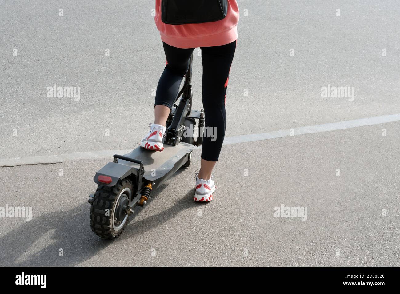 The legs of an unknown girl in white sneakers and gym leggings riding on a black electric scooter over urban asphalt. Modern transportation, electric Stock Photo