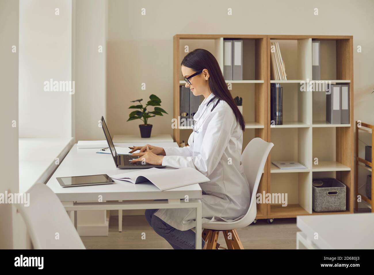 Smiling woman doctor sitting in medical clinic with laptop and typing something during online consultation Stock Photo