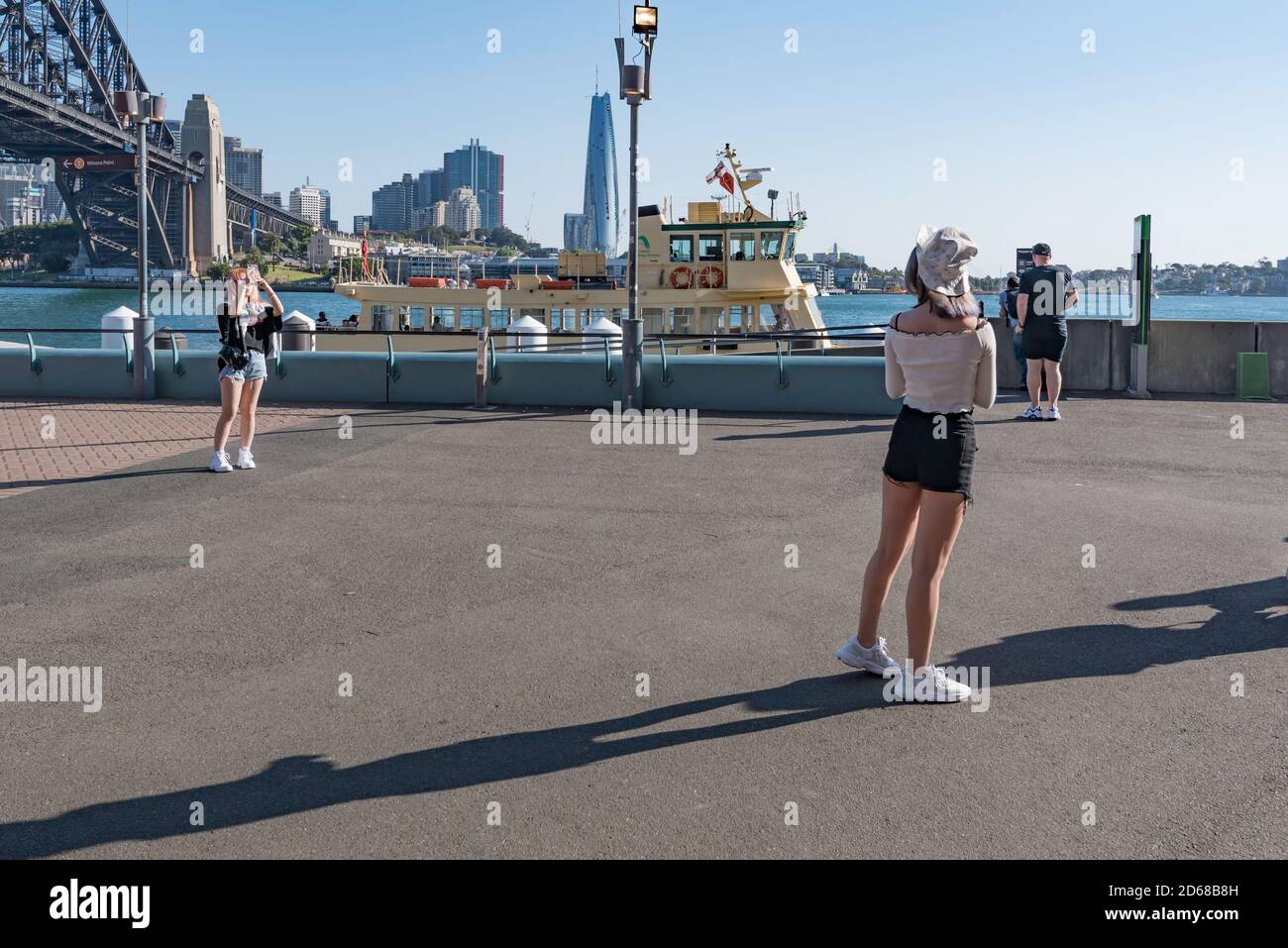 A young Asian woman poses while her friend takes her photo with a mobile phone in front of Luna Park, Sydney, Australia Stock Photo