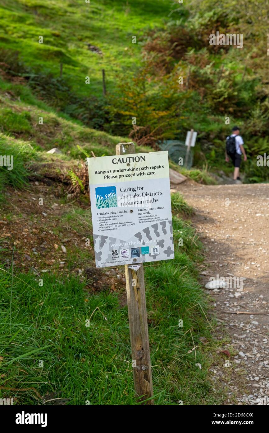 Close up of ‘Fix the fells’ sign beside footpath on Loughrigg Fell with walker man person walking in the background Lake District Cumbria England UK Stock Photo