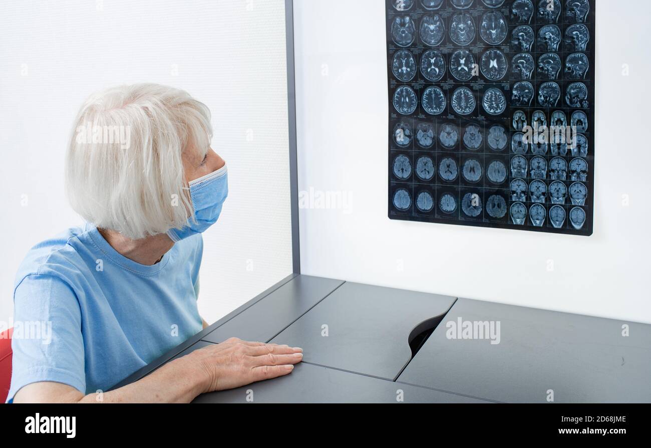 Elderly woman patient wearing a medical mask looks at the results of an MRI scan of her brain. Diagnosis of diseases and head injuries. DIAGNOSTICS OF Stock Photo