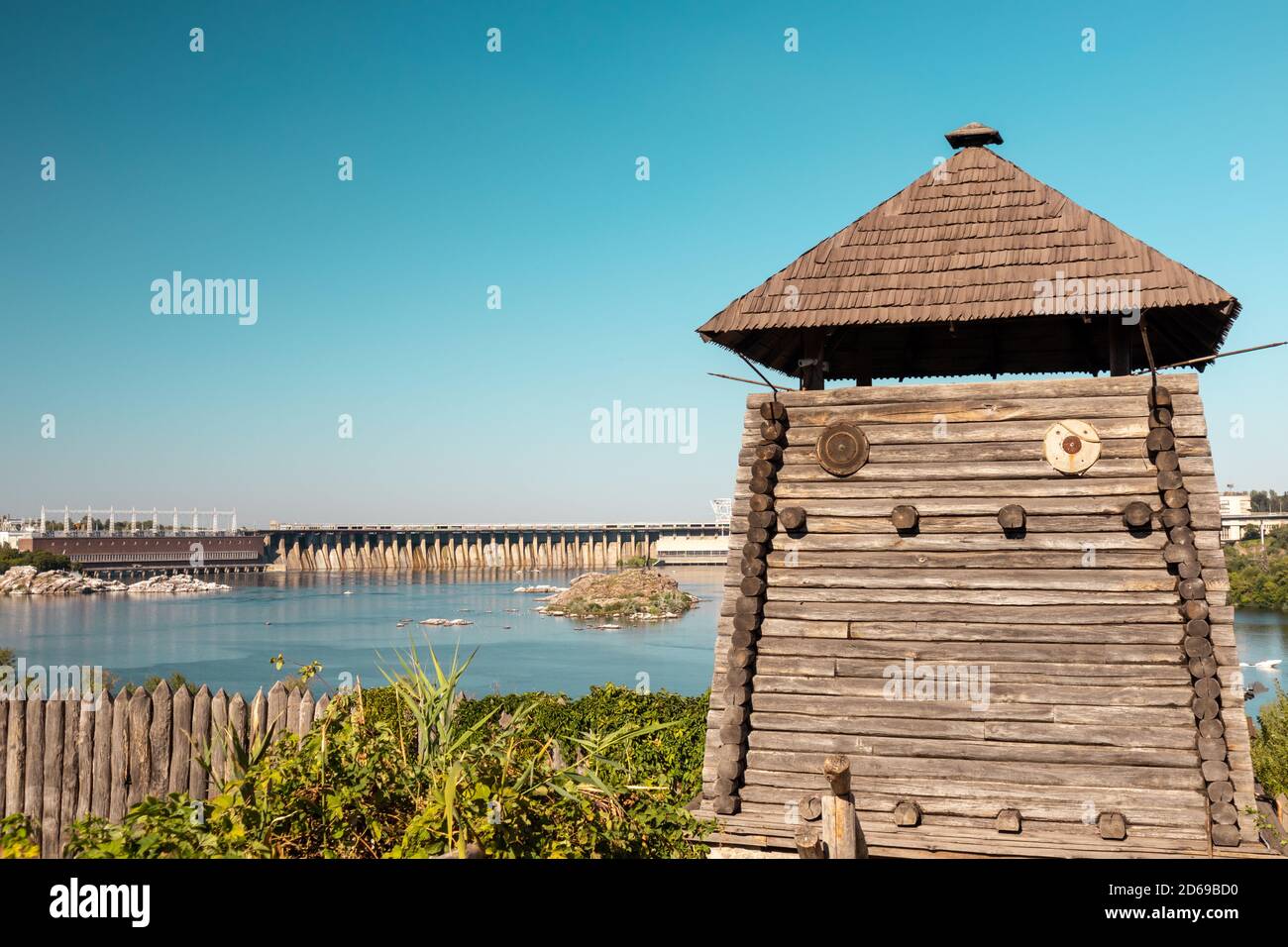 Watchtower in wooden medieval Zaporozhian Sich historical complex, view on DneproGES power station with blue clear bright sky. Khortytsia island on Dn Stock Photo