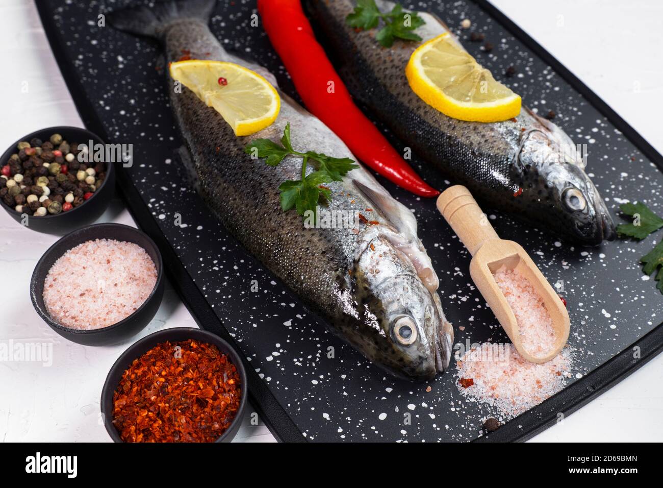 Fresh rainbow trouts on black board on table with salt, pepper, lemon and chili pepper ingredients, Tasty fishes preparing for lunch Stock Photo
