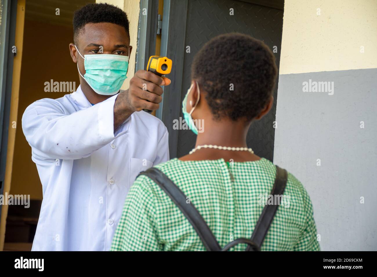 male teacher in an african school checking a pupil's body temperature with an infrared thermometer before she enters the classroom Stock Photo