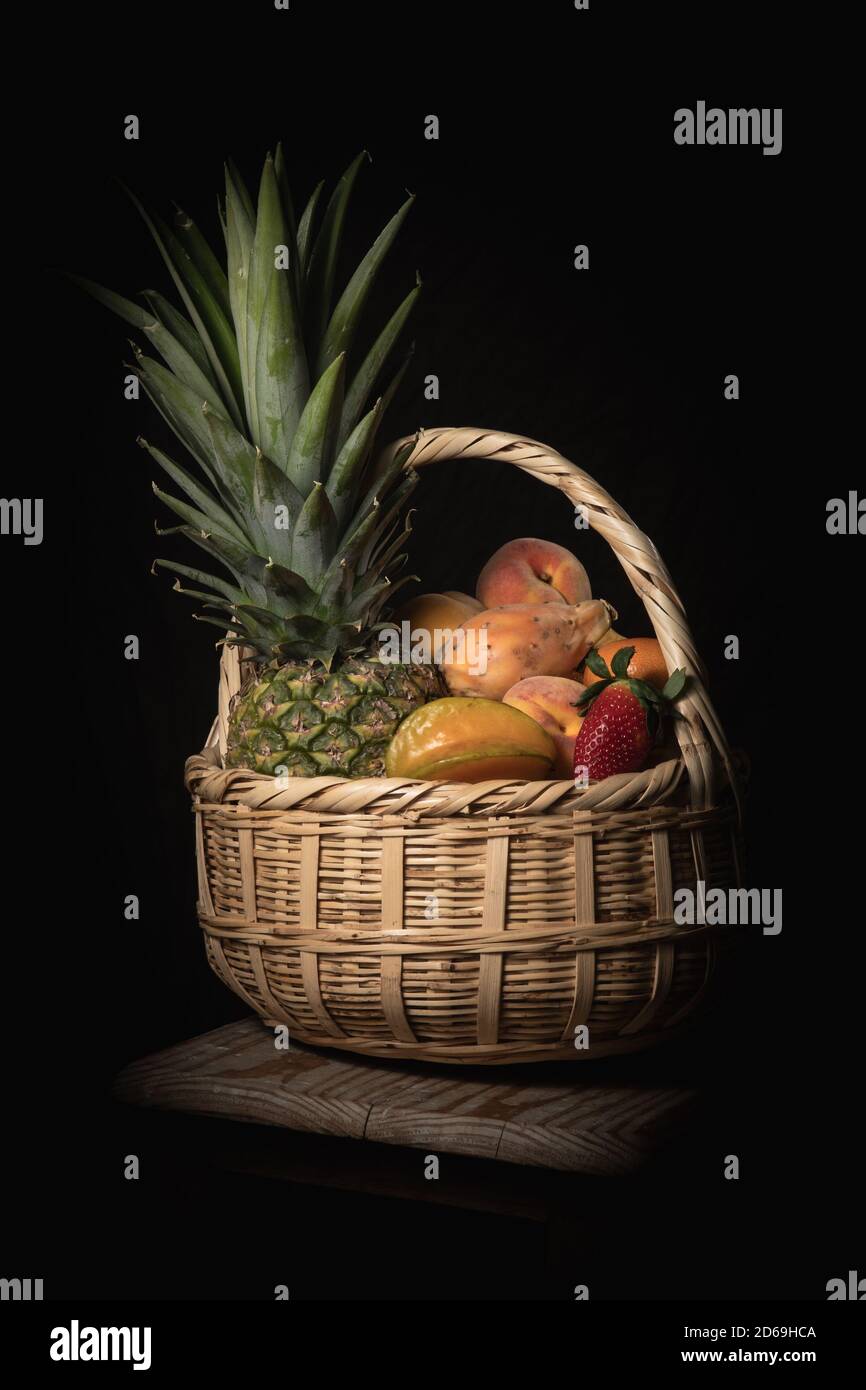 Low-key shot of a tropical fruit basket with pineapple, peach, star fruit, and prickly pear. Stock Photo