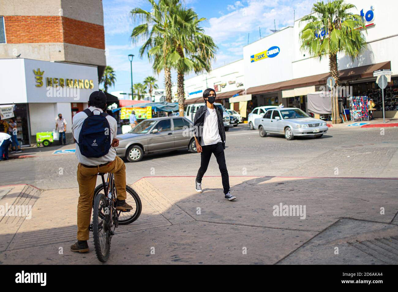 A man rests on his bicycle while a young man walks down the sidewalk  wearing face masks and a pair of converse. Daily life downtown Hermosillo,  Mexico. street photography. © (Photo By