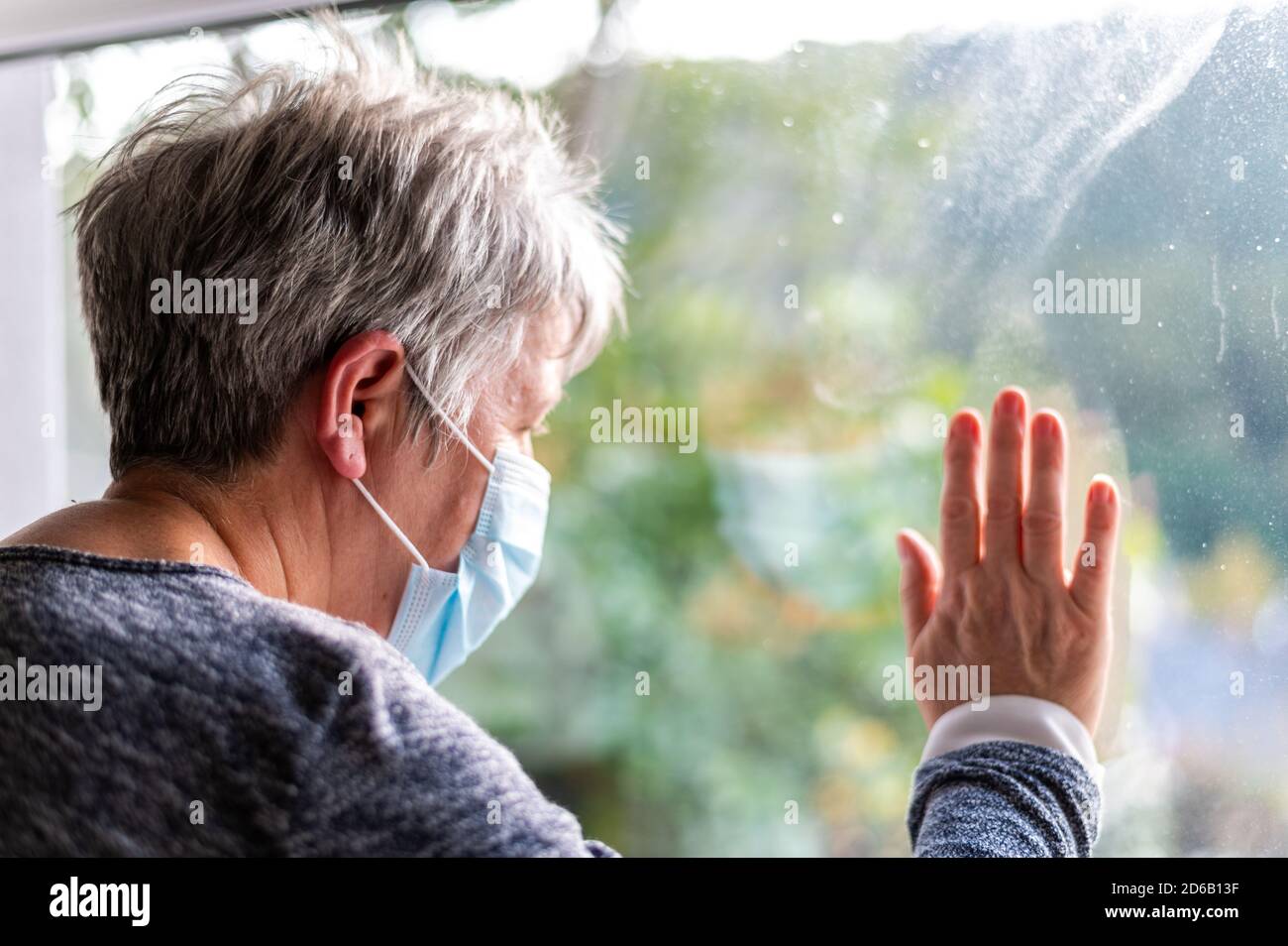 Senior woman wearing protective face mask, looking Out Window, with Covid Virus cells outside of window. Stay Safe stay home concept. shallow focus on Stock Photo