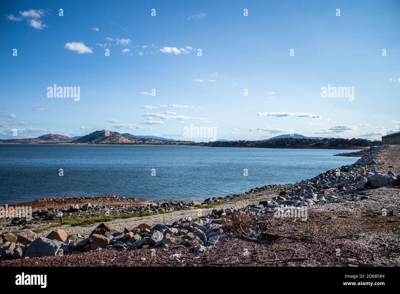 Blue water filled lake dam with rocky foreshore and small hills in background against blue sky Stock Photo