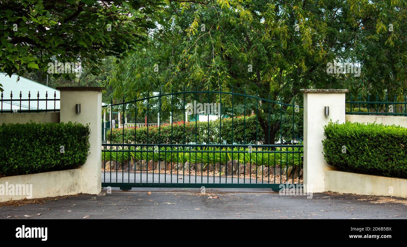 Green metal wrought iron driveway property entrance gates set in concrete  fence, garden shrubs, trees, lights Stock Photo